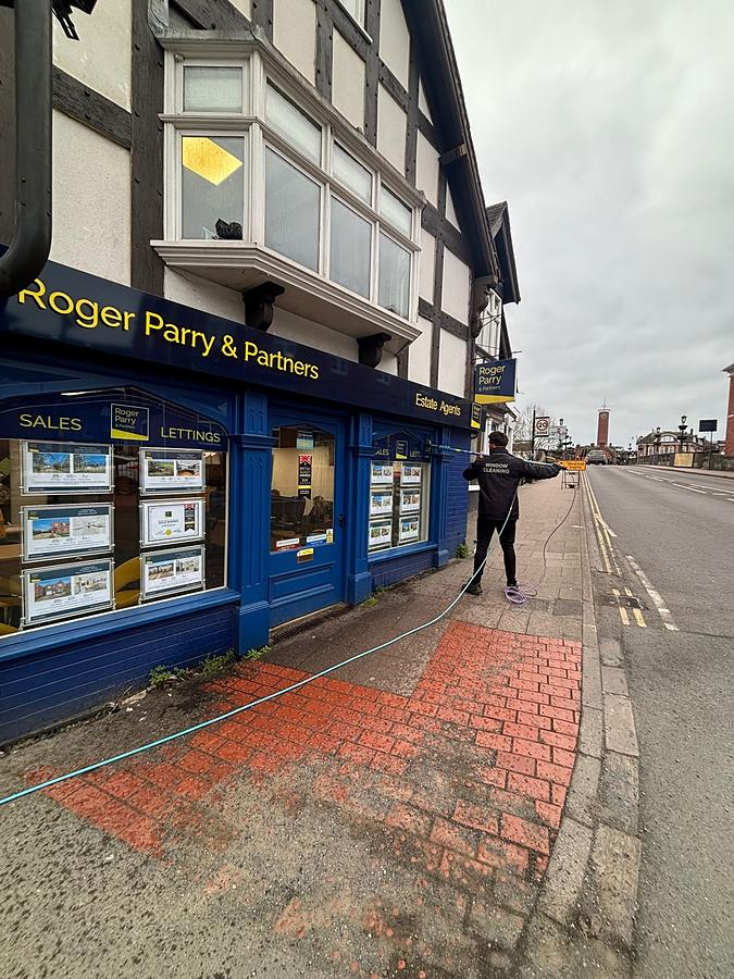 The blue shopfront of Roger Parry and Partners estate agents below a black and white timber framed building, with the brick pavement outside part way through a pressure wash.